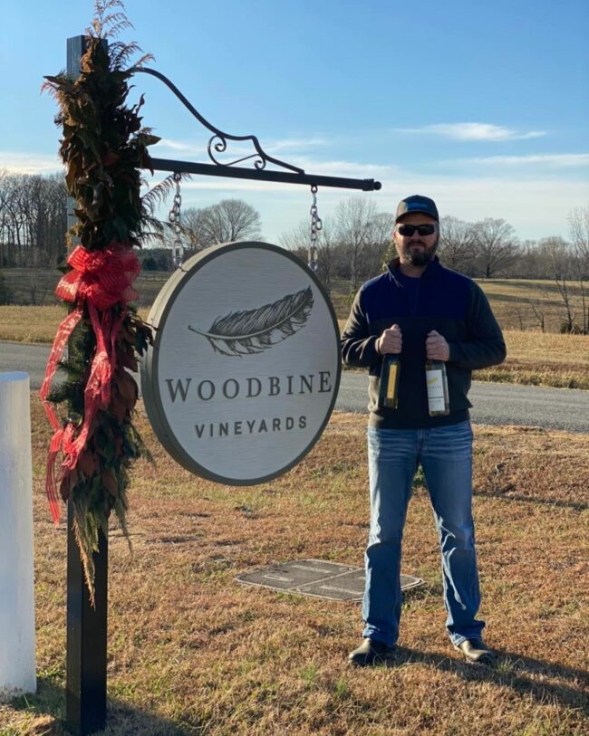 Man holding two bottles of Woodbine Vineyards in front of winery sign